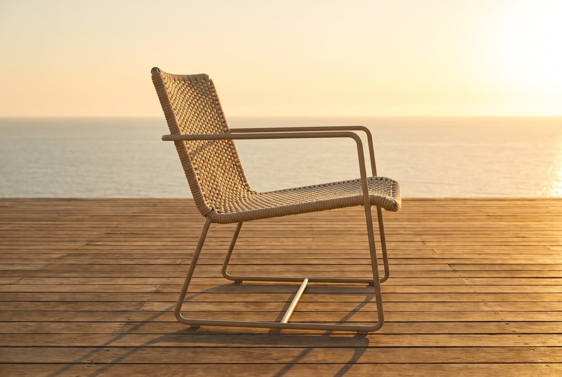 Poltrona de área externa com assento e encosto trançados em corda náutica, sozinha num deck de madeira ao fim da tarde, com o mar ao fundo, imagem de referência.